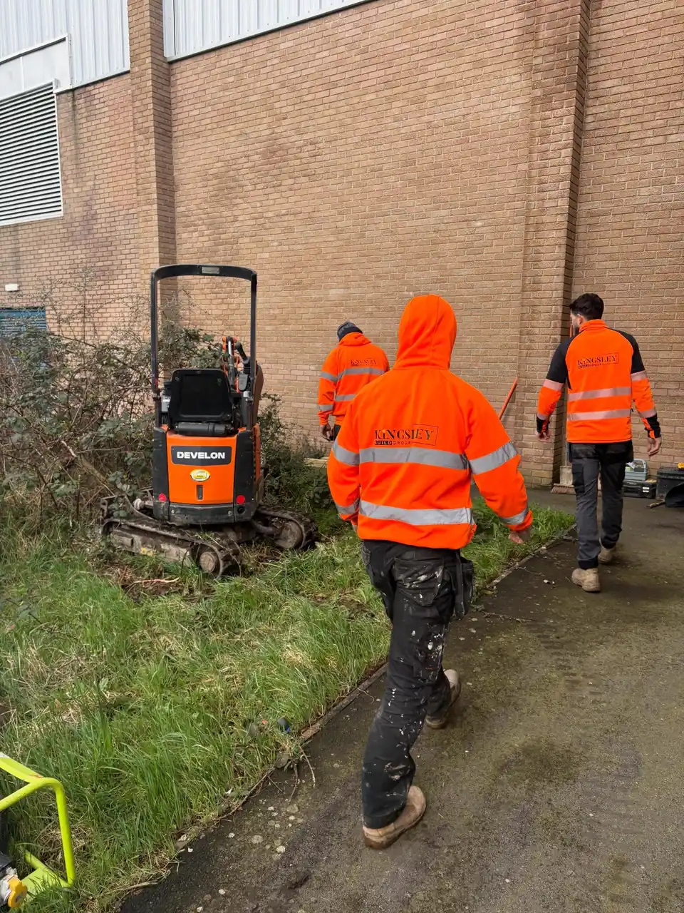 Kingsley team with a mini excavator clearing overgrown ground beside a building